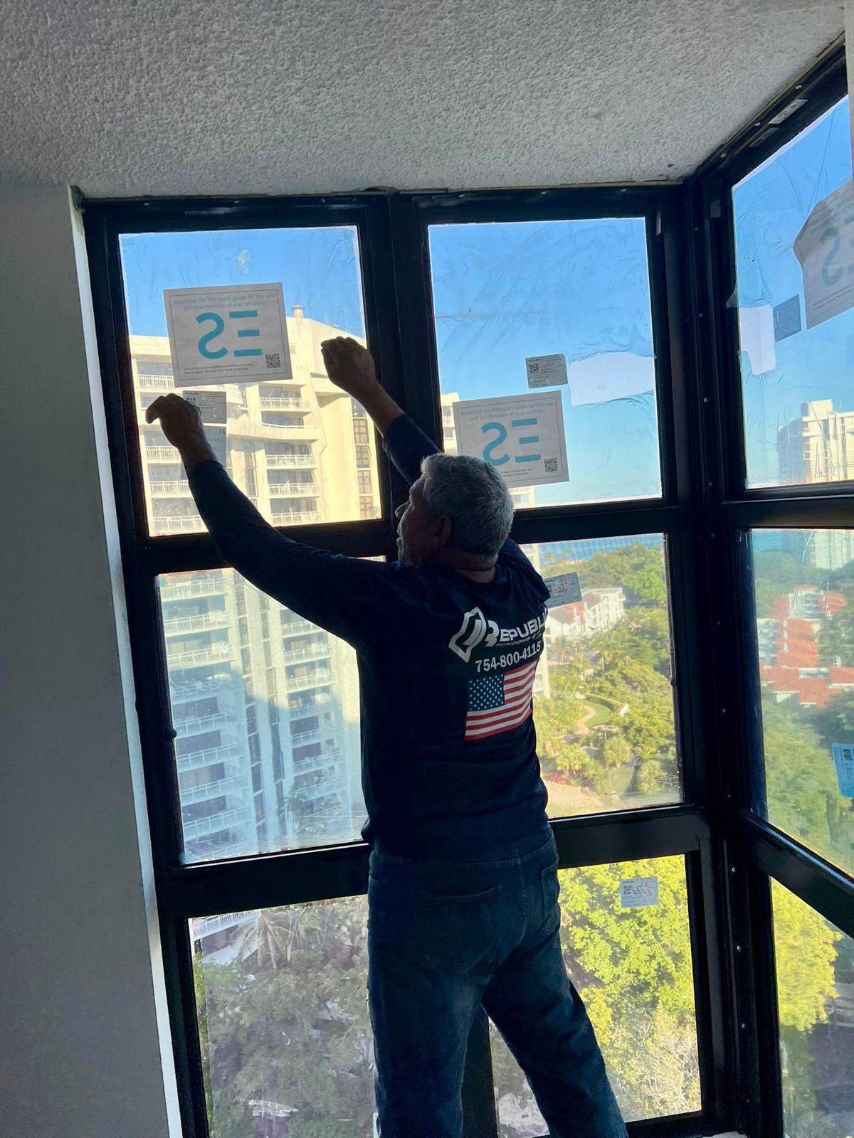 Republic technician in navy uniform installing a dark bronze ES Windows (ESW) multi-lite corner impact window assembly in an upper-floor South Florida high-rise apartment