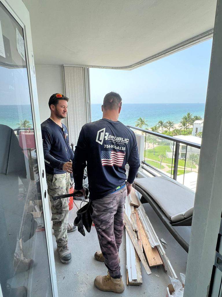 Two Republic Windows & Doors technicians on an upper-floor oceanfront condominium balcony installing heavy-duty impact sliding glass doors overlooking the beach and Atlantic Ocean