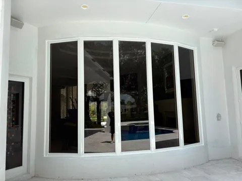 Curved bay of five tall white-frame fixed impact windows on a covered patio, with the pool reflected in the glass