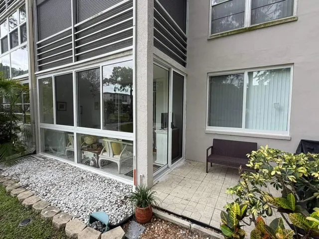 Wide exterior view of a ground-floor Florida room enclosure with white aluminum impact windows and a glass entry door beside rock landscaping