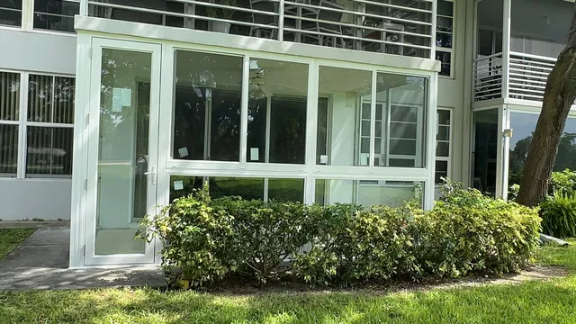Exterior view of a ground-floor patio enclosure with white-frame fixed picture windows, horizontal rollers, and a single impact glass door