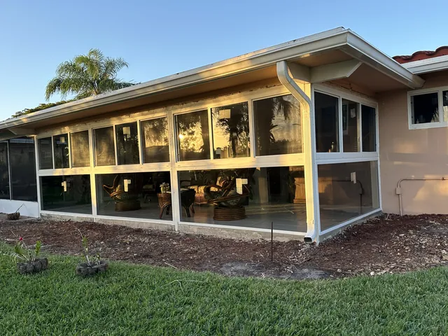 Wide exterior view of a single-story Florida room enclosure with white-frame impact windows under an asphalt shingle roofline
