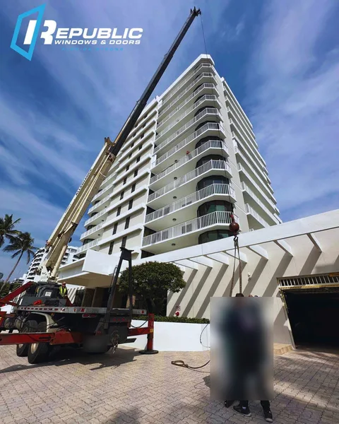 Crane truck lifting a crated impact glass unit up the side of an oceanfront condo tower while a Republic installer guides it