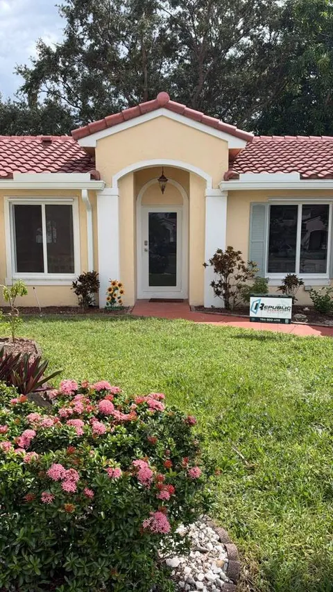 Tan stucco home with red clay tiles, white impact windows, an arched entry, and a Republic yard sign by pink flowers
