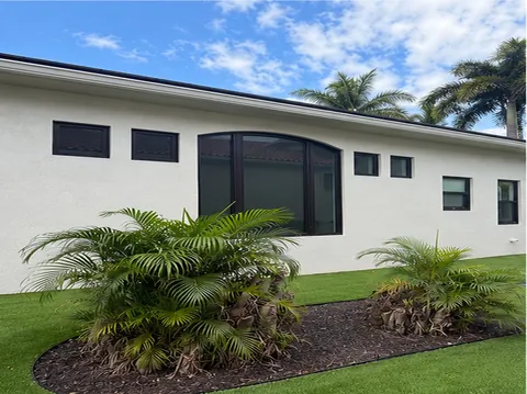 White home with a large dark-frame arched impact window and small square windows behind palm shrubs