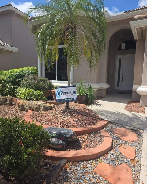 White-frame impact window and entry door behind a palm, with a Republic yard sign in the bed